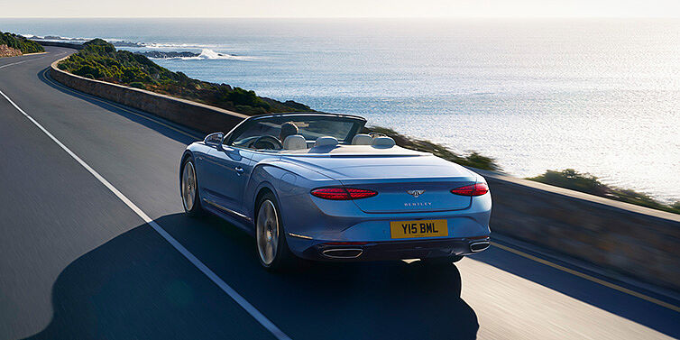 Bentley Basel New Bentley Continental GTC Azure convertible rear view in Light Windsor blue paint with 22 inch ten spoke wheels driving dynamically on an ocean road in the sunshine