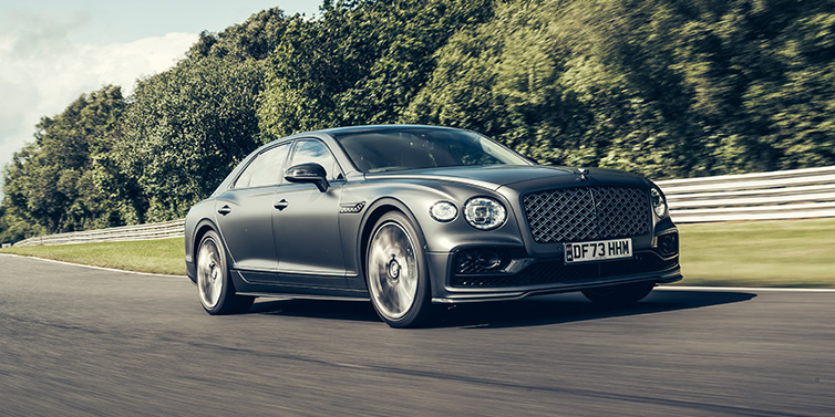 Bentley Basel Bentley Flying Spur Mulliner sedan front three quarter in dark grey paint driving dynamically by some trees on a track