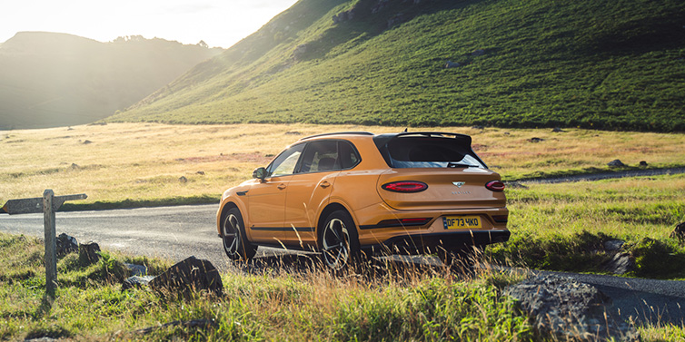 Bentley Basel Bentley Bentayga S SUV rear three quarter in Sunburst Gold paint on a road surrounded by green hills