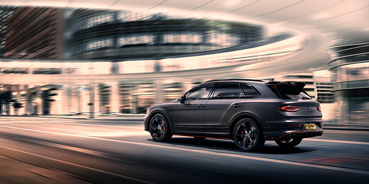 Bentley Basel Bentley Bentayga S Black Edition SUV rear three quarter in Anthracite Satin paint driving dynamically through a city at night