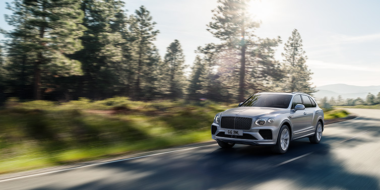 Bentley Basel Bentley Bentayga SUV front three quarter in Moonbeam silver paint driving dynamically along a tree-lined road