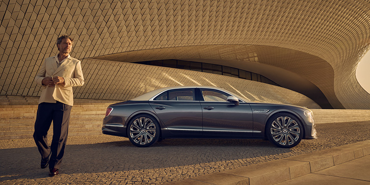 Bentley Basel Bentley Flying Spur Mulliner sedan in Rose Gold over Magnetic duotone paint parked by a curved wall with person in suit jacket standing on the left